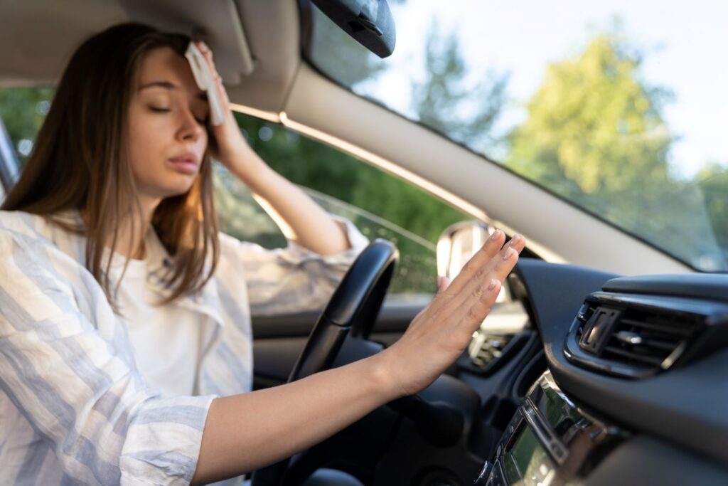 female driver wiping sweat with a hand on the ac vent inside a car